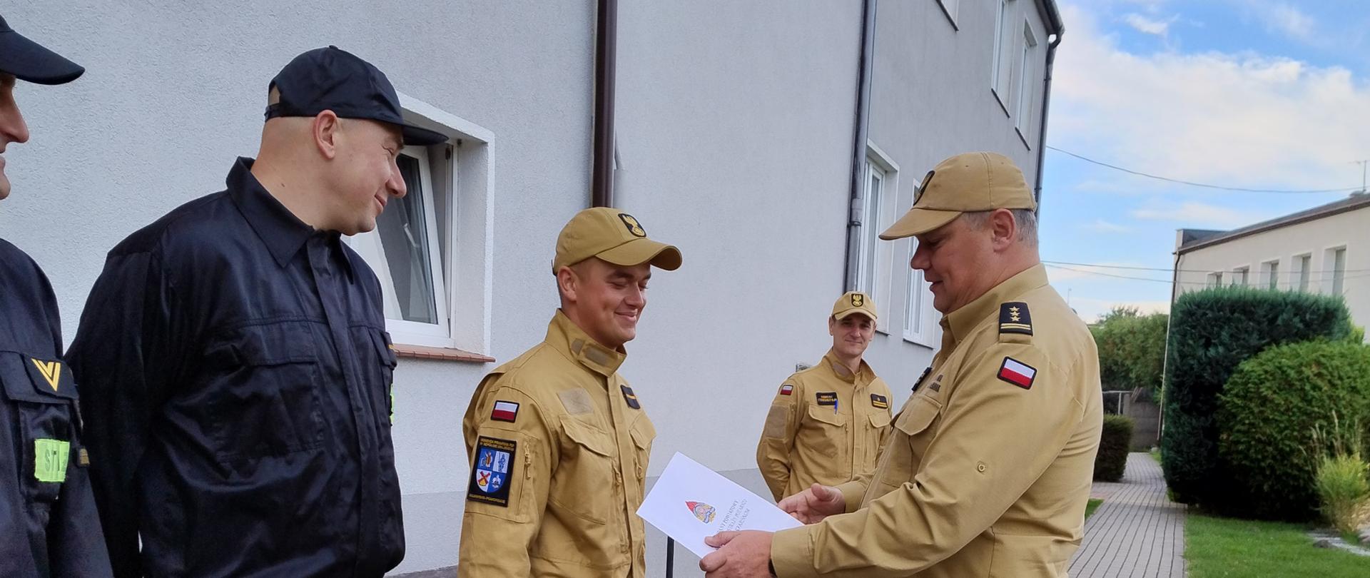 Kadr zrobiony z bocznej perspektywy przedstawia moment gratulacji na placu przed strażnicą. Po prawej stronie stoi dowódca w piaskowym mundurze i czapce, trzymający biały dyplom, który podaje rękę młodemu strażakowi w identycznym, jasnym stroju. Po lewej stronie, w rzędzie, stoją pozostali strażacy w czarnych kombinezonach koszarowych – wszyscy patrzą na ceremonię i uśmiechają się. W tle widać fragment czerwonej bramy garażowej, chodnik, zielony trawnik oraz ozdobne krzewy.