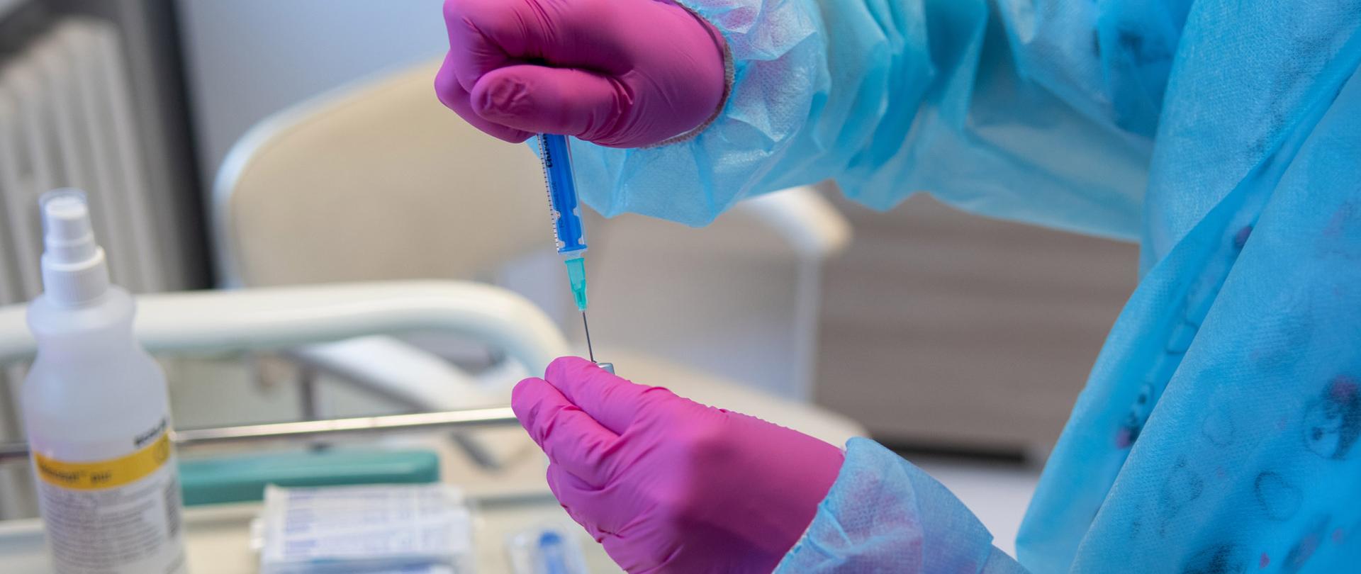 Na zdjęciu dłonie w rękawicach ochronnych a w nich nabierana do strzykawki szczepionka przeciwko koronawirusowi. 