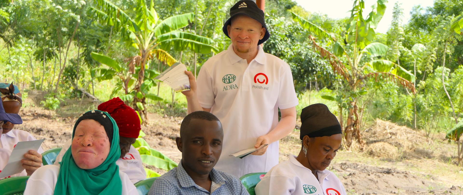 Grupa osób, część z koszulkach z napisame Adra/Polish Aid siedzi na krzesłach, dwie osoby są Albinosami