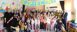 Na zdjęciu widać wszystkich uczestników kampanii prewencyjnej o nazwie Zawsze uważny zawsze bezpieczny w przedszkolu w pionkach. Nad uczestnikami wisi żółty napis na niebieskim tle o nazwie kampanii.