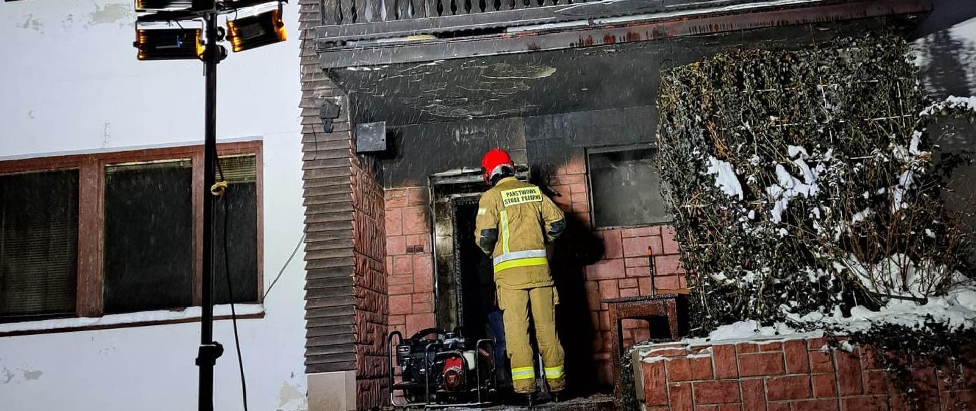 Budynek mieszkalny, piętrowy. Przed drzwiami wejściowymi stoi strażak w umundurowaniu bojowym. W oknach widać spalone wnętrze obiektu.