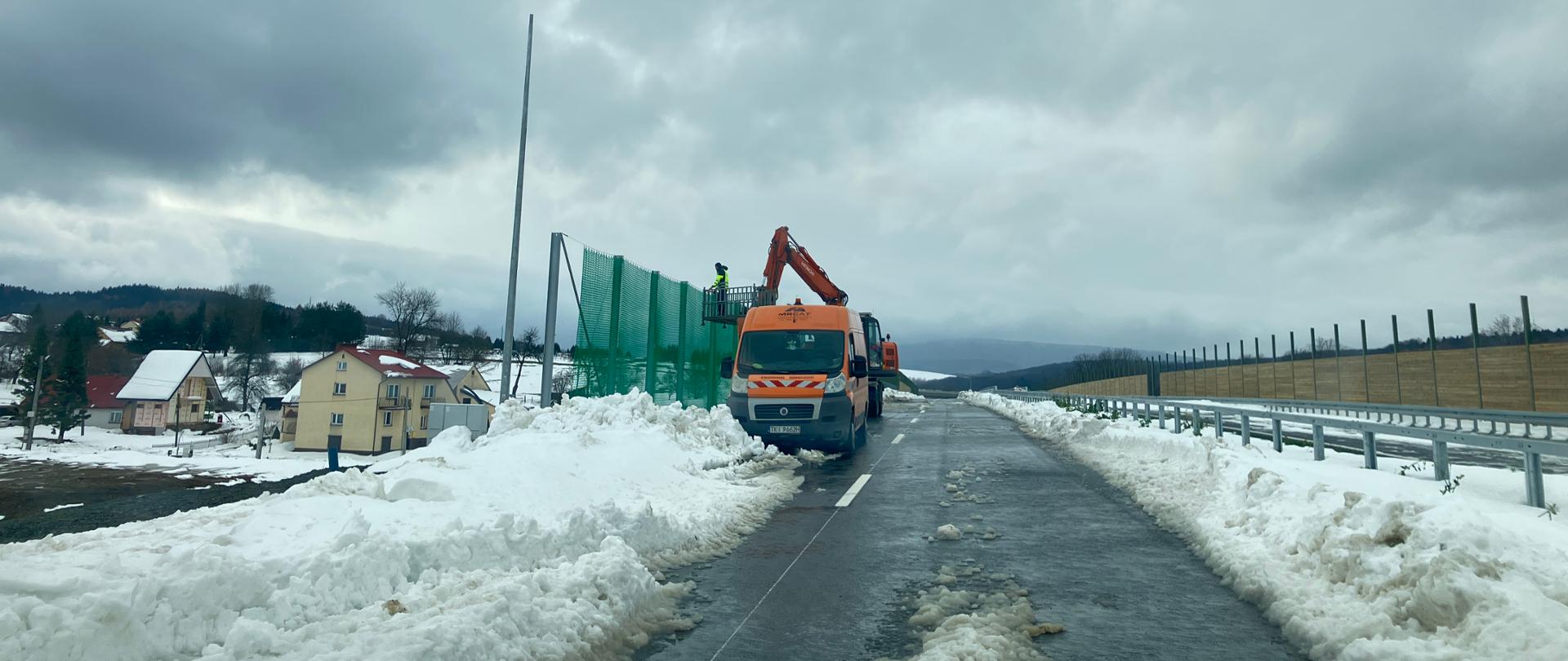 Droga w zimowej scenerii, częściowo odśnieżona, z mokrym śniegiem na nawierzchni. Po obu stronach wysokie zaspy. Na środku stoi pomarańczowa ciężarówka z wysięgnikiem, obok montowane są zielone ekrany akustyczne. W tle domy z ośnieżonymi dachami i pagórkowaty teren. Niebo całkowicie zachmurzone.