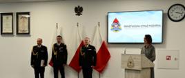 Wojewoda pomorska stoi i przemawia przy mównicy za nią stoi kadra kierownicza Państwowej Straży Pożarnej ustawione są flagi Polski na ścianie wisi rota ślubowania ekran z logiem i napisem Państwowej Straży Pożarnej oraz zegar.
