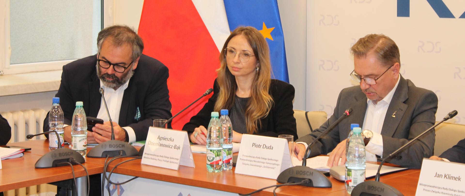 Na zdjęciu widnieje stół prezydialny za którym siedzą od lewej strony: Liwiusz Laska – Dyrektor Generalny MRPiPS , Agnieszka Dziemianowicz-Bąk Ministra Rodziny, Pracy i Polityki Społecznej, Wiceprzewodnicząca Rady Dialogu Społecznego, Piotr Duda – przewodniczący Rady Dialogu Społecznego. W tle siedzących dwie flagi: UE i Polski.