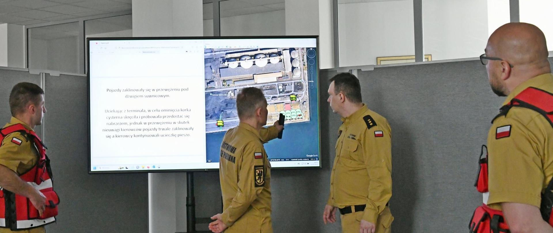 Dwóch funkcjonariuszy stoi przy monitorze na którym wyświetlany jest zakres ćwiczenia dwóch strażaków w czerwonych kamizelkach obserwuje ich działania.