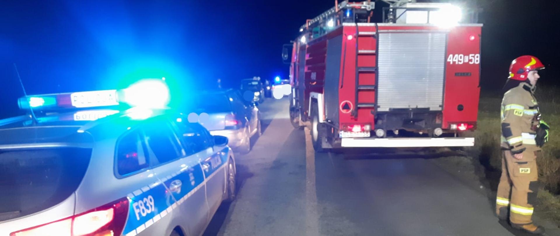 Na zdjęciu widać czerwony samochód straży pożarnej. Z jego lewej strony stoi radiowóz policyjny i ciemny samochód osobowy. Z tyłu samochodu stoi strażak w żółtym stroju i czerwonym hełmie. Przed tymi samochodami w oddali stoją dwa następne samochody osobowe. Wszystkie pojazdy stoją na drodze asfaltowej. Jest ciemno.