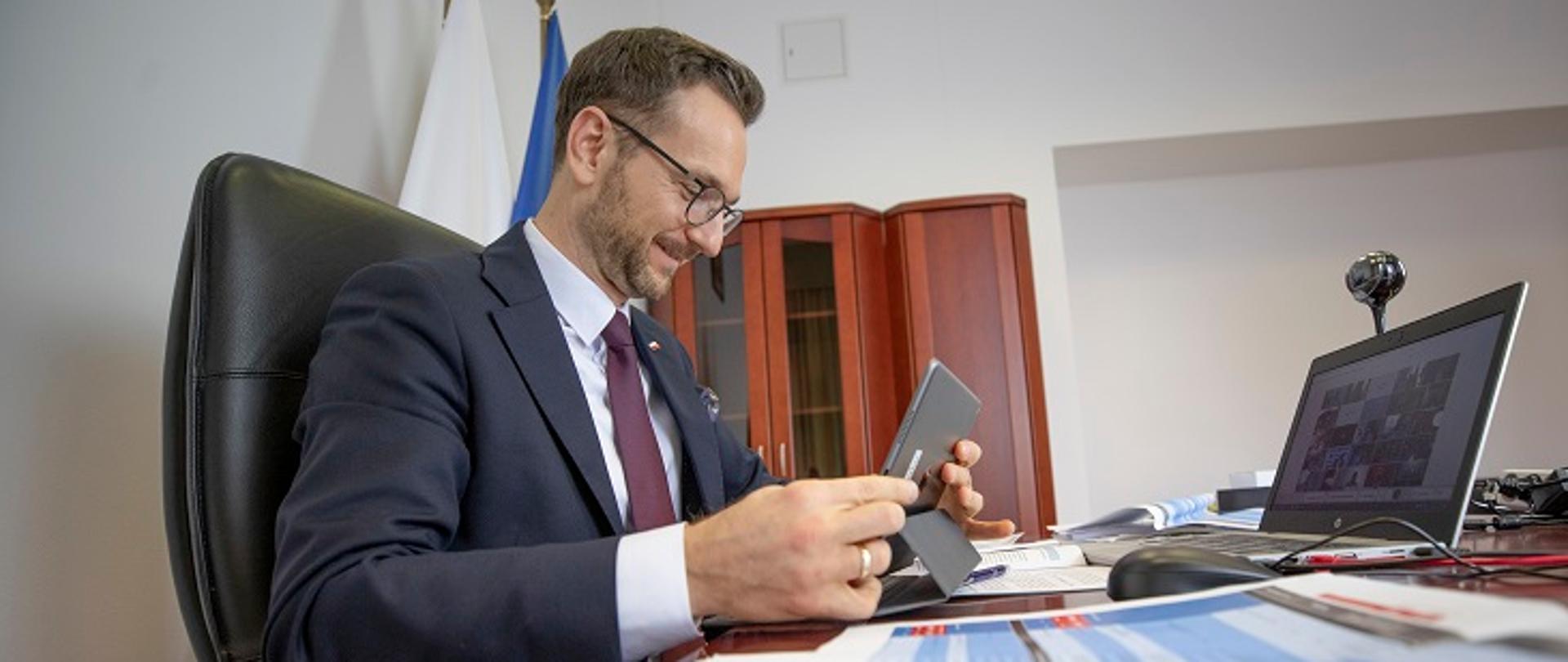 Wiceminister Waldemar Buda siedzi przy biurku, na którym stoi laptop i leżą dokumenty. Minister trzyma w dłoniach tablet.