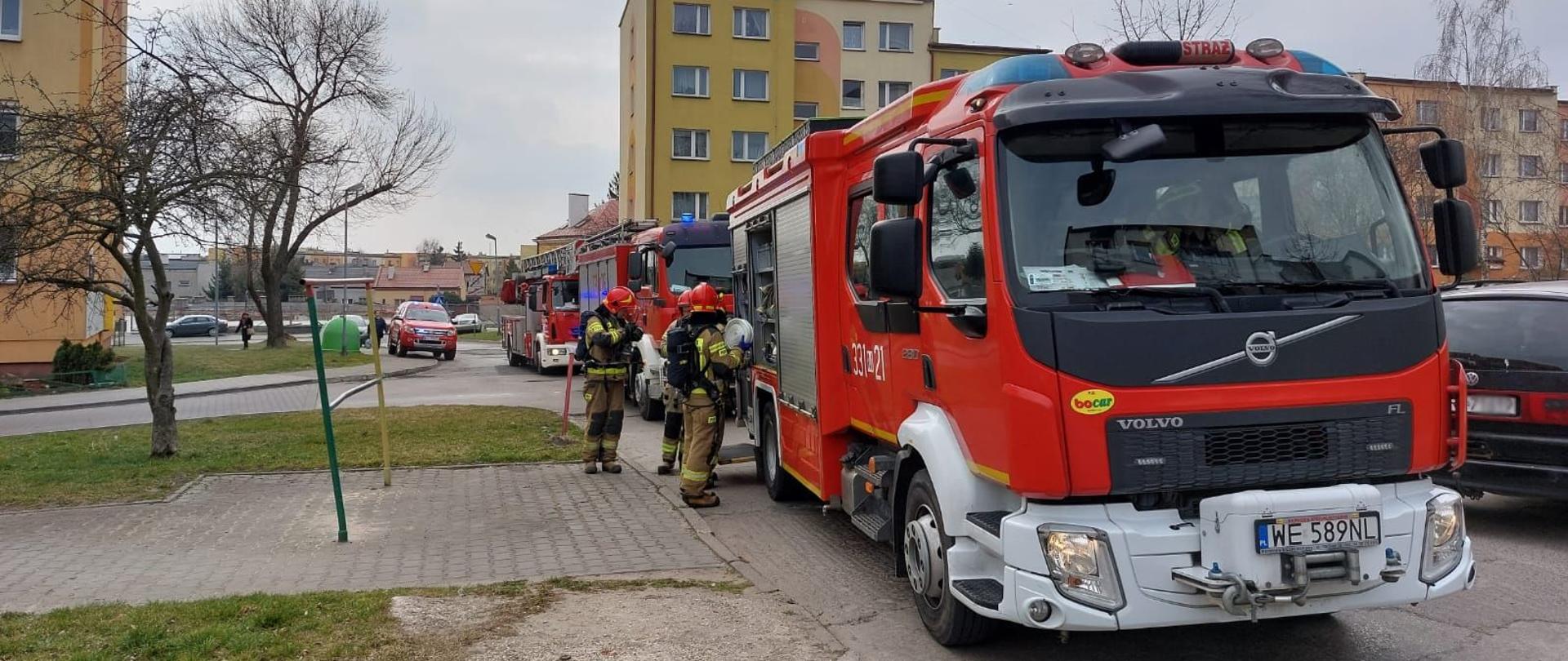 Zdjęcie wykonane przed budynkiem mieszkalnym w którym wybuch pożar. Przed budynkiem widać 4 samochody pożarnicze ustawione jeden za drugim. Pojazdy w kolorze czerwonym z białymi i srebrnymi elementami zabudowy. Dwa pierwsze to pojazdy ratowniczo – gaśnicze posiadające między innymi zbiorniki z wodą do celów gaśniczych. Trzecim pojazdem jest pojazd z drabiną mechaniczną umożliwiający dotarcie ratownikom na wyższe piętra. Ostatni z nich to pojazd terenowy typu pick-up. Pojazd przeznaczony jest dla dowódcy jednostki ratowniczo – gaśniczej. Przed pierwszymi pojazdami znajdują się strażacy. Ubrani są w ubrania specjalne w kolorze piaskowym. Na głowach maja pozakładane czerwone hełmy. Kilku z nich na plecach ma założony sprzęt ochrony dróg oddechowych w postaci butlowych aparatów ze sprężonym powietrzem. Dodatkowo jeden z nich trzyma zwinięty w krąg wąż pożarniczy.