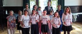 Zdjęcie grupowe uczennic i nauczycieli. Uczestnicy zdjęcia stoją w 3 rzędach. Wszyscy wykonawcy trzymają na wysokości klatki piersiowej biały dyplom. W tle widać kolorowe witrażowe okna oraz fragment fortepianu.