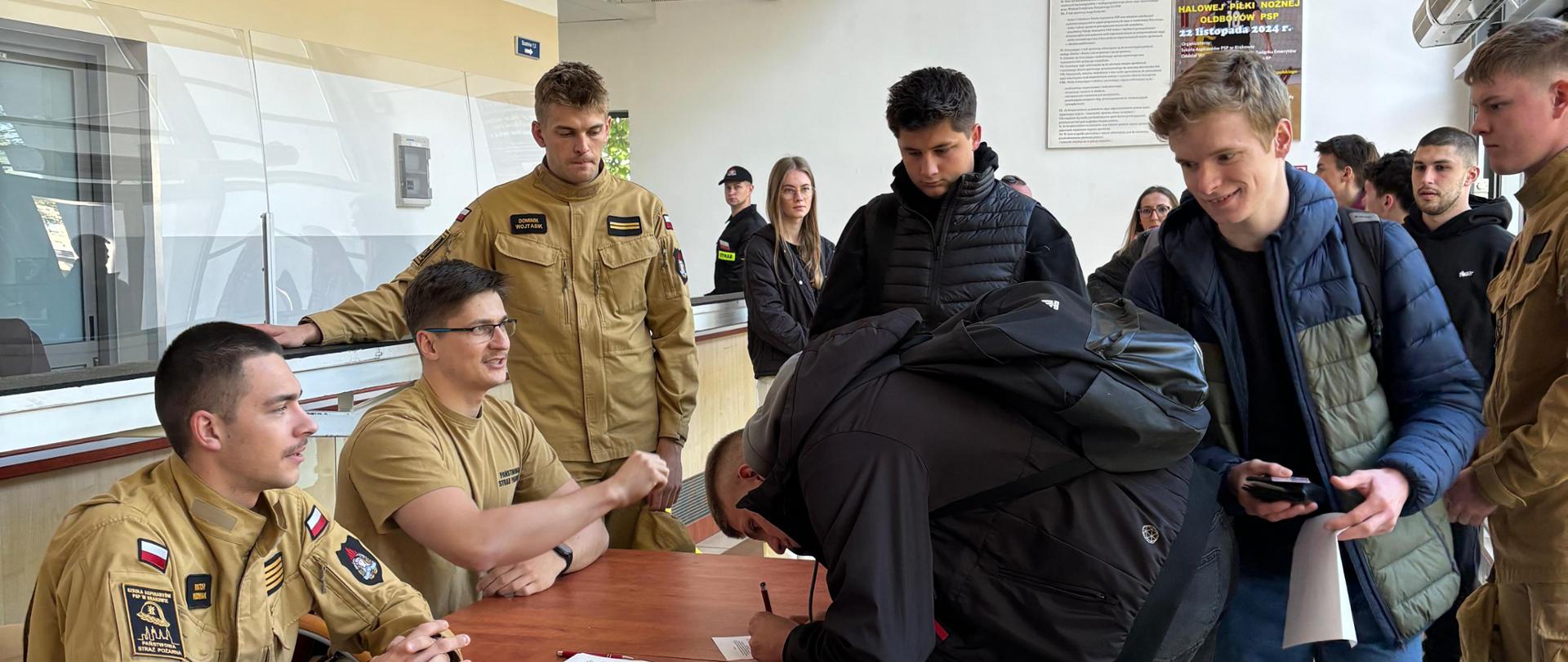 Zdjęcie wykonane w przedsionku sali gimnastycznej. W centralnej części zdjęcia drewniany stolik przy którym siedzi 2 i stoi 1 funkcjonariusz PSP w piaskowych ubraniach służbowych. przedsionek wypełniony jest osobami w codziennych ubraniach cywilnych. Jeden z nich pochyla się nad stolikiem i pisze na białej kartce. Pomieszczenie wysokie jasnooświetlone, w górnej części widoczne szaliki różnych klubów piłkarskich.