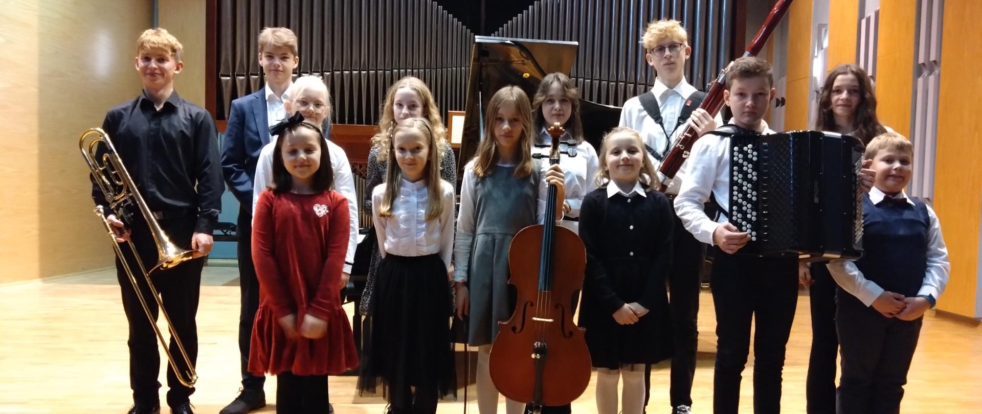 Na scenie sali koncertowej w dwóch rzędach stoją dzieci i trzymają w dłoniach swoje instrumenty. Za nimi fortepian a w tle organy.