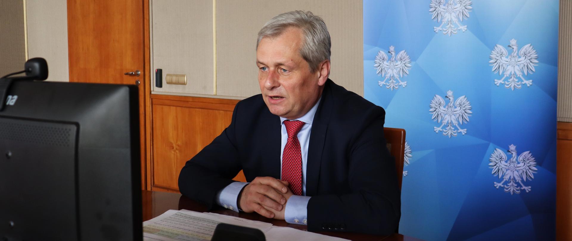 Na zdjęciu siedzący za stołem, wojewoda opolski Sławomir Kłosowski. Mężczyzna siedzi nieco pochylony nad stołem, patrzy wprost - tj. w monitor komputera. Łokcie ma oparte o stół. Jest w trakcie rozmowy. Przed nim na stole lezą kartki, notatki. W tle za jego plecami widać granatowy roll up z napisem wojewoda opolski. Po lewej stronie kadru widać fragment monitora komputerowego oraz głośnik. 