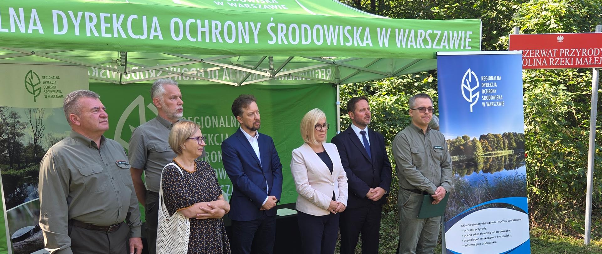 Na zdjęciu przedstawiono grupę uczestników wydarzenia zorganizowanego na świeżym powietrzu, w otoczeniu lasu i zieleni. Osoby pozują do wspólnego zdjęcia przed zielonym namiotem oraz banerami Regionalnej Dyrekcji Ochrony Środowiska w Warszawie. W tle widoczna jest również tablica oznaczająca Rezerwat Przyrody Dolina Rzeki Małej. Całość odbywa się w słoneczny dzień, a leśne otoczenie i materiały informacyjne podkreślają przyrodniczy oraz edukacyjny charakter wydarzenia.