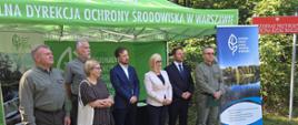 Na zdjęciu przedstawiono grupę uczestników wydarzenia zorganizowanego na świeżym powietrzu, w otoczeniu lasu i zieleni. Osoby pozują do wspólnego zdjęcia przed zielonym namiotem oraz banerami Regionalnej Dyrekcji Ochrony Środowiska w Warszawie. W tle widoczna jest również tablica oznaczająca Rezerwat Przyrody Dolina Rzeki Małej. Całość odbywa się w słoneczny dzień, a leśne otoczenie i materiały informacyjne podkreślają przyrodniczy oraz edukacyjny charakter wydarzenia.