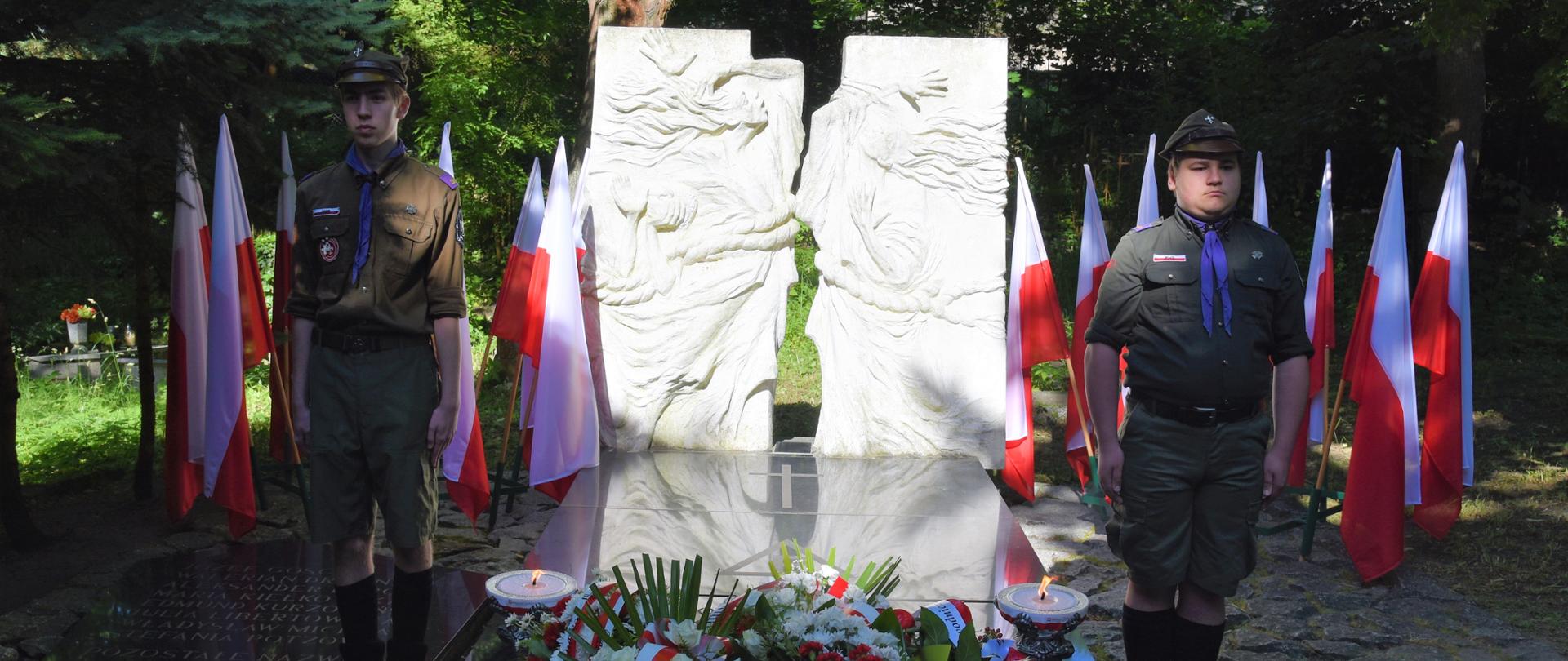 Przy pomniku wartę trzyma dwóch harcerzy. Na płycie płoną dwa znicze i leżą wiazanki kwiatów. 