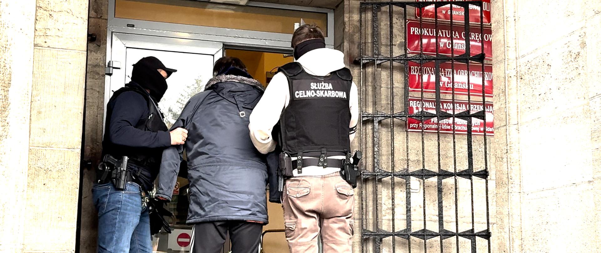 Trzech mężczyzn stoi przy drzwiach wejściowych budynku; dwóch w kamizelkach służbowych eskortuje osobę w ciemnej kurtce, obok widoczne czerwone tablice urzędowe.
