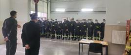 Zdjęcie przedstawia garaże Komendy Powiatowej Państwowej Straży Pożarnej, w których odbywa się podsumowanie szkolenia podstawowego strażaków ratowników OSP. Na tle garażu strażacy OSP, zastępca Komendanta Powiatowego oraz przedstawiciel Komendanta Wojewódzkiego PSP..
