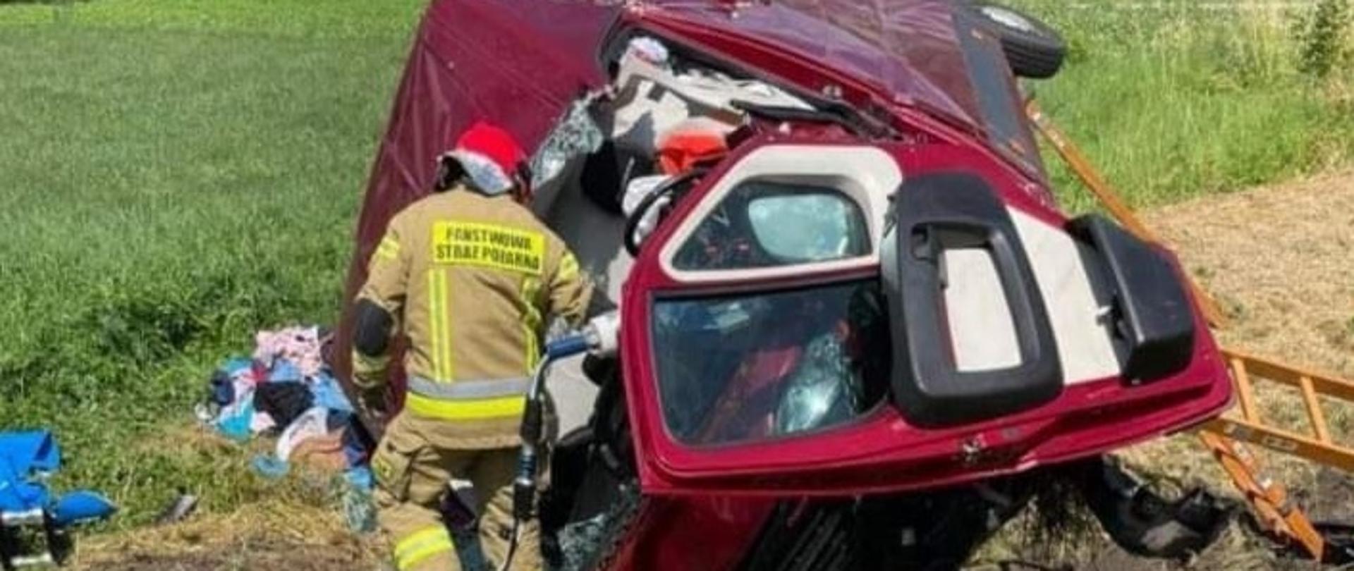 Zdjęcie przedstawia czerwony samochód dostawczy, który brał udział w zdarzeniu. Obok samochodu stoi strażak pracujący sprzętem hydraulicznym.
