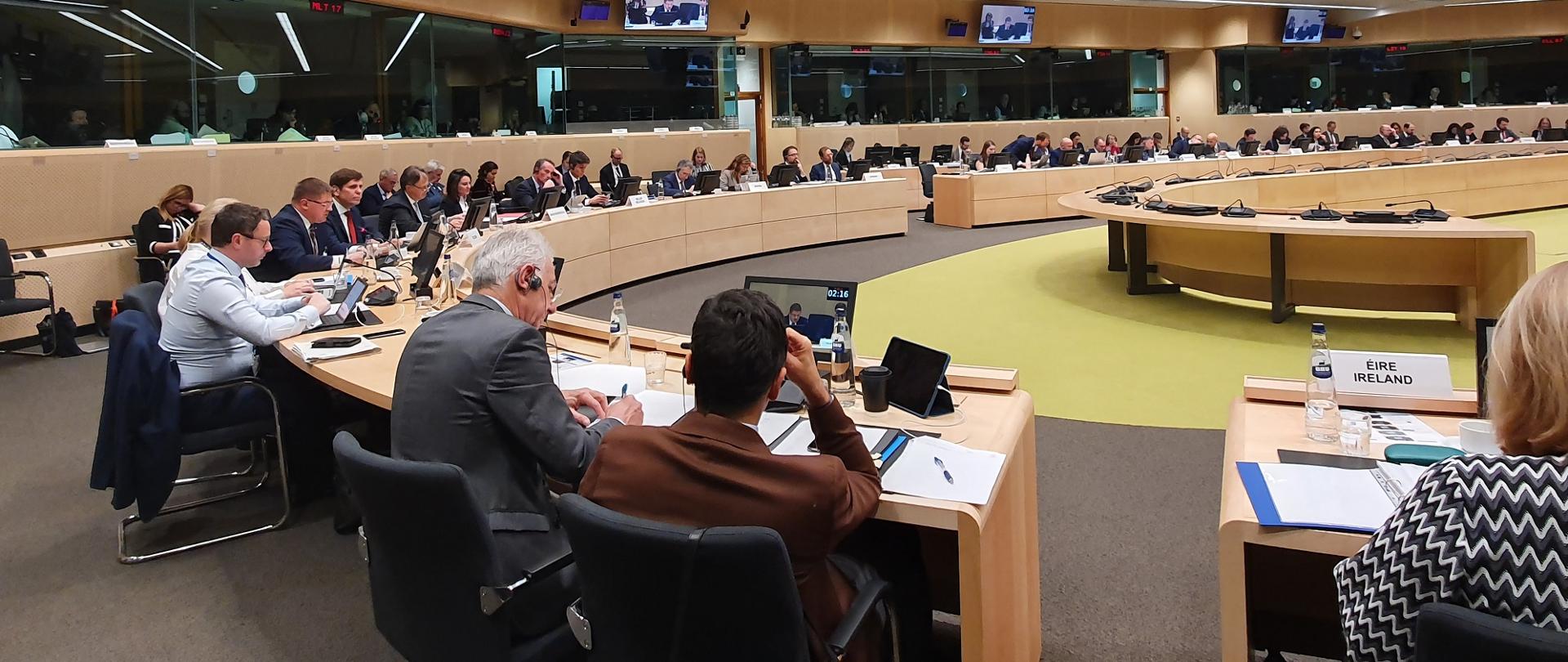 Widok na dużą salę, przy stołach ustawionych w okrąg siedzą ludzie, za nimi szklane ściany.