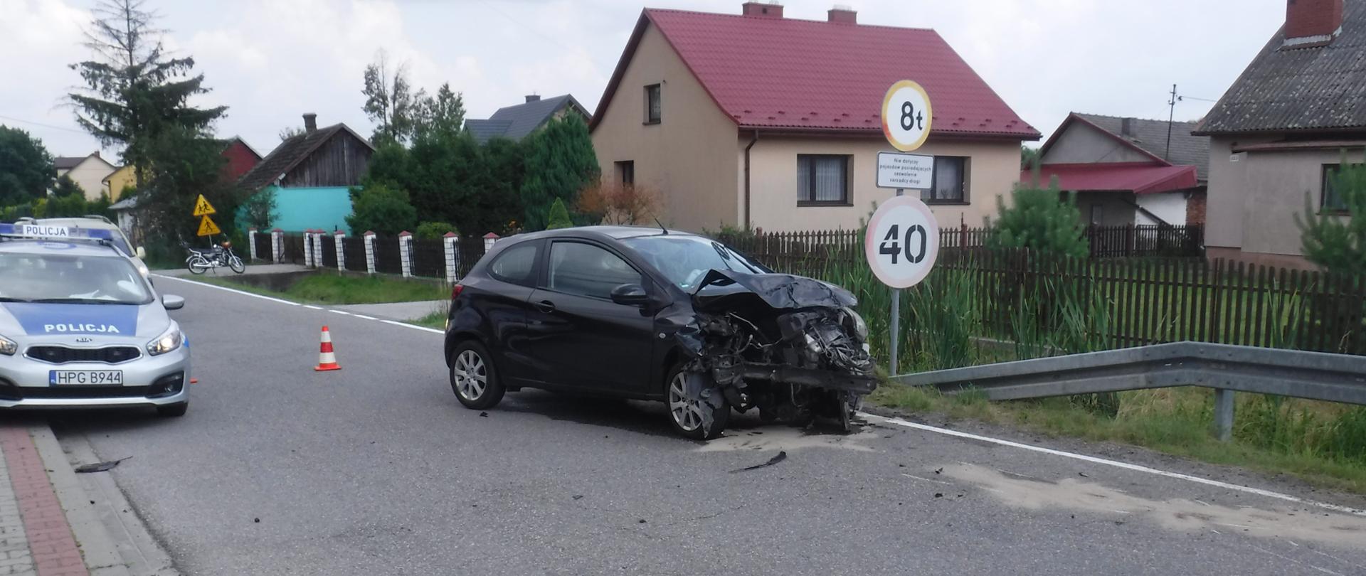 Zdjęcie główne przedstawia samochód osobowy koloru czarnego. Posiada on zgniecioną całą komorę silnika. Jezdnia wokół samochodu jest posypana sorbentem w celu zebrania substancji ropopochodnych, które w trakcie zdarzenia wyciekły z pojazdu. Po lewej stronie zdjęcia widać radiowóz policji. Po prawe stronie widać bariery energochłonne wraz z osoba postronną ubraną w kamizelkę odblaskową.