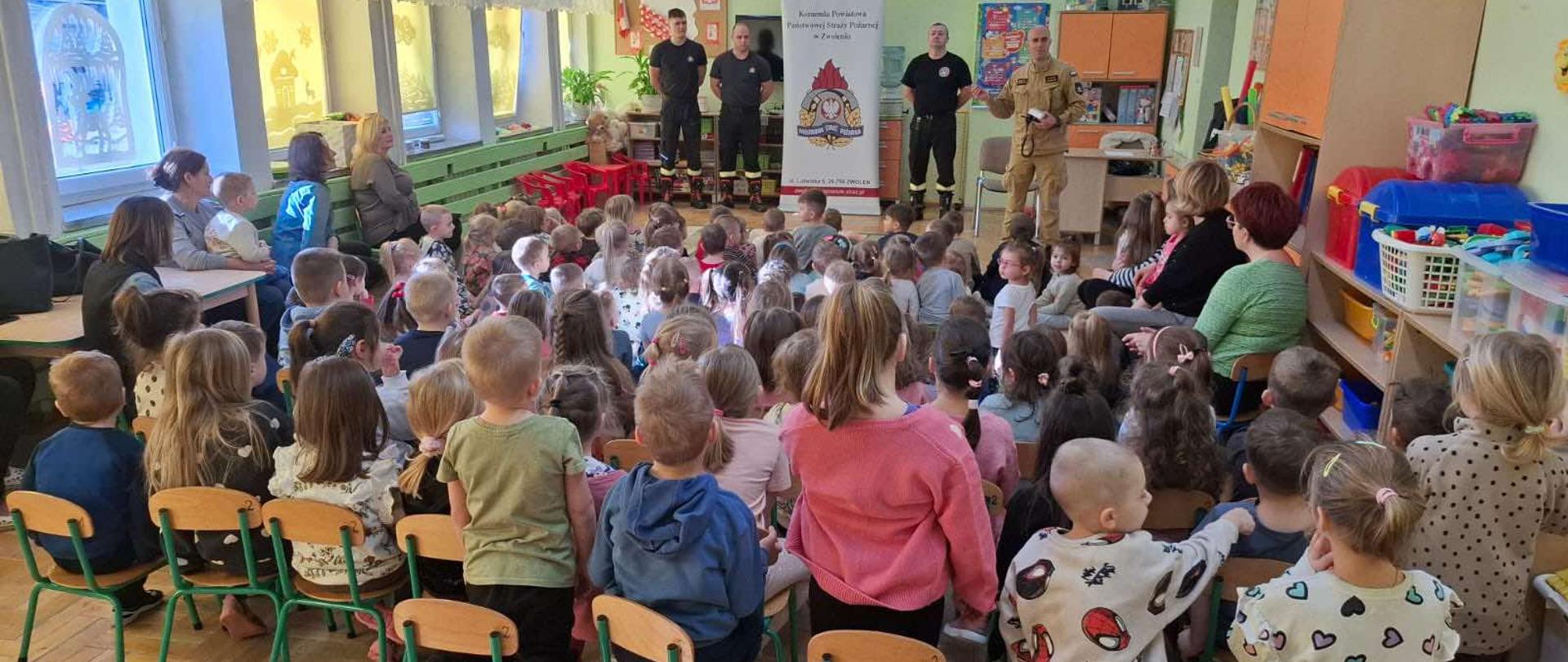 Zdjęcie przedstawia spotkanie lub zajęcia edukacyjne w przedszkolu, z udziałem przedstawicieli Państwowej Straży Pożarnej w Zwoleniu. Grupa dzieci siedzi na małych krzesłach, zwrócona w stronę dorosłych, którzy stoją z przodu sali. Na środku stoi banner z logo Komendy Powiatowej Państwowej Straży Pożarnej w Zwoleniu. Kilku dorosłych w mundurach strażackich lub roboczych prezentuje coś dzieciom. Pomieszczenie jest jasno oświetlone i ozdobione, typowo dla przedszkola, z zabawkami i materiałami edukacyjnymi widocznymi w tle.