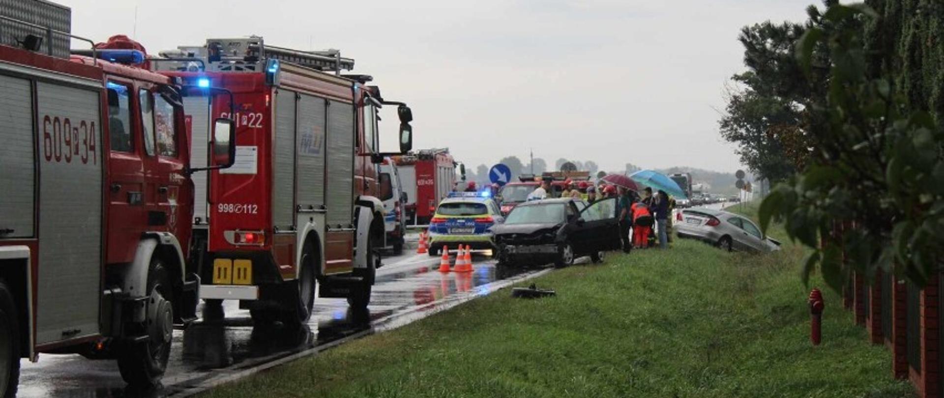 Na zdjęciu czerwone samochody strażackie oraz karetka pogotowia ratunkowego, w tyle strażacy wraz z ratownikami medycznymi udzielający pomocy poszkodowanym, przed nimi wrak pojazdu
