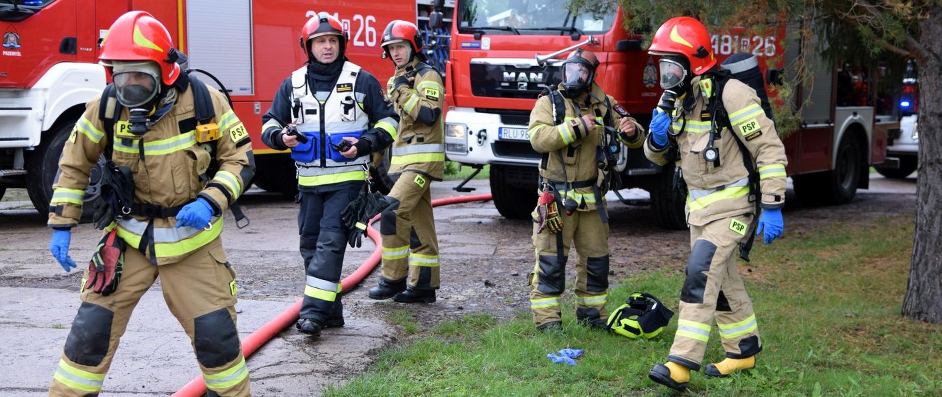 Zdjęcie zrobione na zewnątrz, podczas ćwiczeń. Pięciu strażaków ubrany w ubrania specjalne przygotowuje się do podjęcia działań ratowniczo-gaśniczych. W tle samochody ratowniczo-gaśnicze