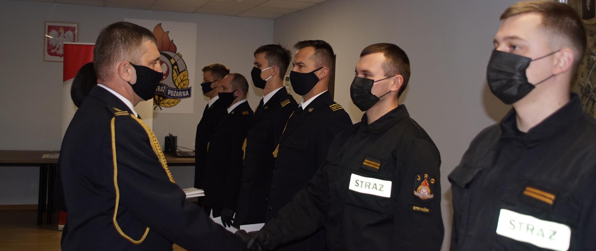 Komendant Powiatowy PSP w Zgierzu st. bryg. Paweł Malinowski wręcza awanse strażakom którzy znajdują się na sali konferencyjnej w Komedzie Powiatowej Państwowej Straży Pożarnej w Zgierzu. Strażacy stoją przy jasnoszarej ścianie na której wisi obraz z historią pożarnictwa. Odznaczani funkcjonariusze przedstawieni są w strojach wyjściowych oraz koszarowych.