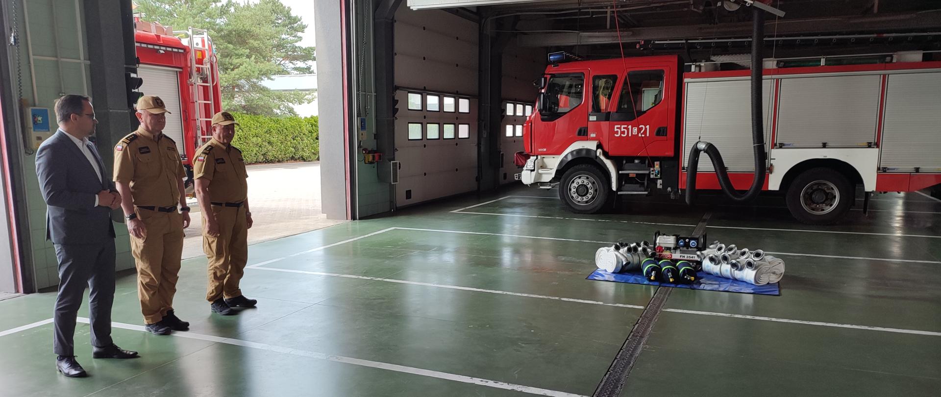Dwóch strażaków w umundurowaniu musztardowym stoi obok mężczyzny w garniturze obok na niebieskiej płachcie leży sprzęt a niedaleko stoi samochód gaśniczy.