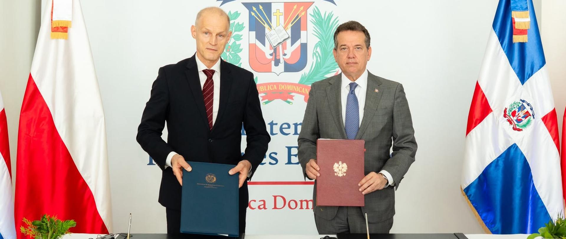 Two diplomats stand behind a table during a document-signing ceremony. Wojciech Zajączkowski, Under-Secretary of State at the Ministry of Foreign Affairst, wearing a black suit and burgundy tie, holds a navy-blue folder, while Víctor Bisonó Haza, Minister for Foreign Affairs of the Dominican Republic, wearing a grey suit and blue tie, presents a burgundy folder featuring the Polish coat of arms. The flags of Poland and the Dominican Republic, along with the Dominican Republic’s coat of arms, are visible in the background.
