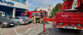Na zdjęciu parking przed budynkiem administracyjno-produkcyjnym firmy DrukPak. Na parkingu znajdują się pojazdy osobowe oraz dwa pojazdy ratownicze należące do Państwowej Straży Pożarnej.
Jeden z nich stojący bliżej budynku to pojazd średni ratowniczo gaśniczy natomiast drugi to pojazd specjalny wyposażony w podnośnik hydrauliczny. W rejonie pojazdów zgromadzeni są strażacy przygotowujący sprzęt do prowadzenia działań ratowniczo-gaśniczych .