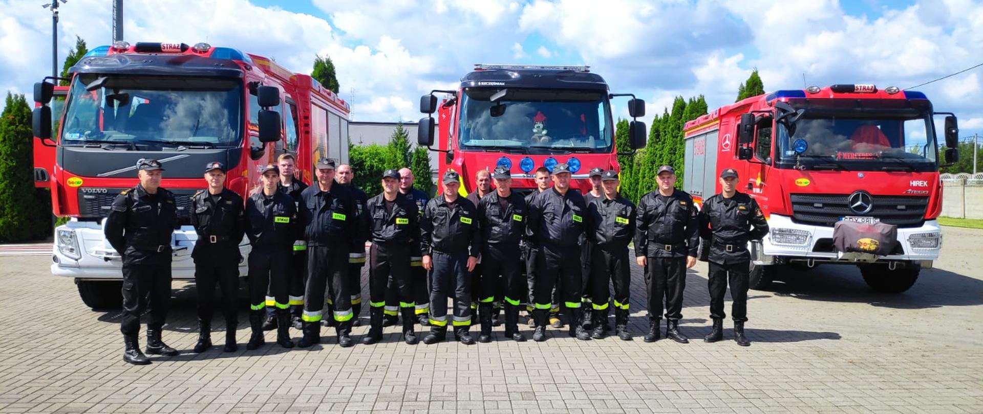 Zdjęcie przedstawia grupę strażaków pozujących do zdjęcia na tle 3 wozów pożarniczych. Zdjęcie wykonane jest na placu, na dworze. 