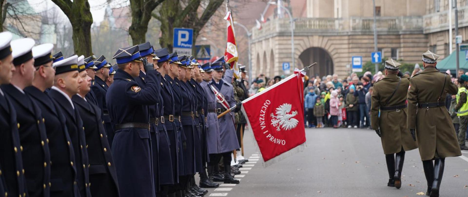Przegląd wojsk i funkcjonariuszy. Od lewej końcówka formacji marynarki wojennej, następnie państwowa straż pożarna, a następnie widoczne sztandary policji itd. W tle cywile oraz widok na budynek Muzeum Narodowego oraz Akademii Morskiej
