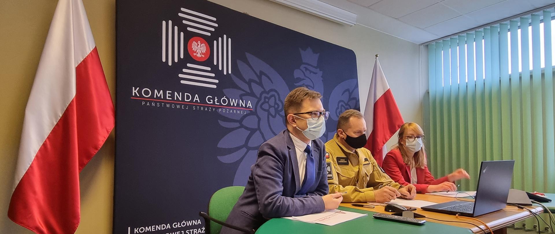 Uczestnicy spotkania w maseczkach ochronnych na tle flag państwowych oraz ścianki z logiem Komendy Głównej Państwowej Straży Pożarnej