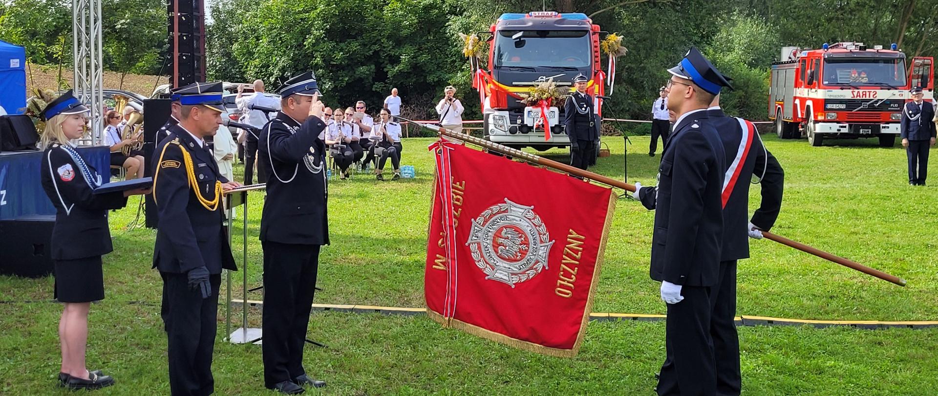 Poczet sztandarowy w trakcie dekoracji sztandaru podczas uroczystej zbiórki. W tle czerwone strażackie samochody.