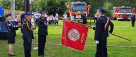 Poczet sztandarowy w trakcie dekoracji sztandaru podczas uroczystej zbiórki. W tle czerwone strażackie samochody.