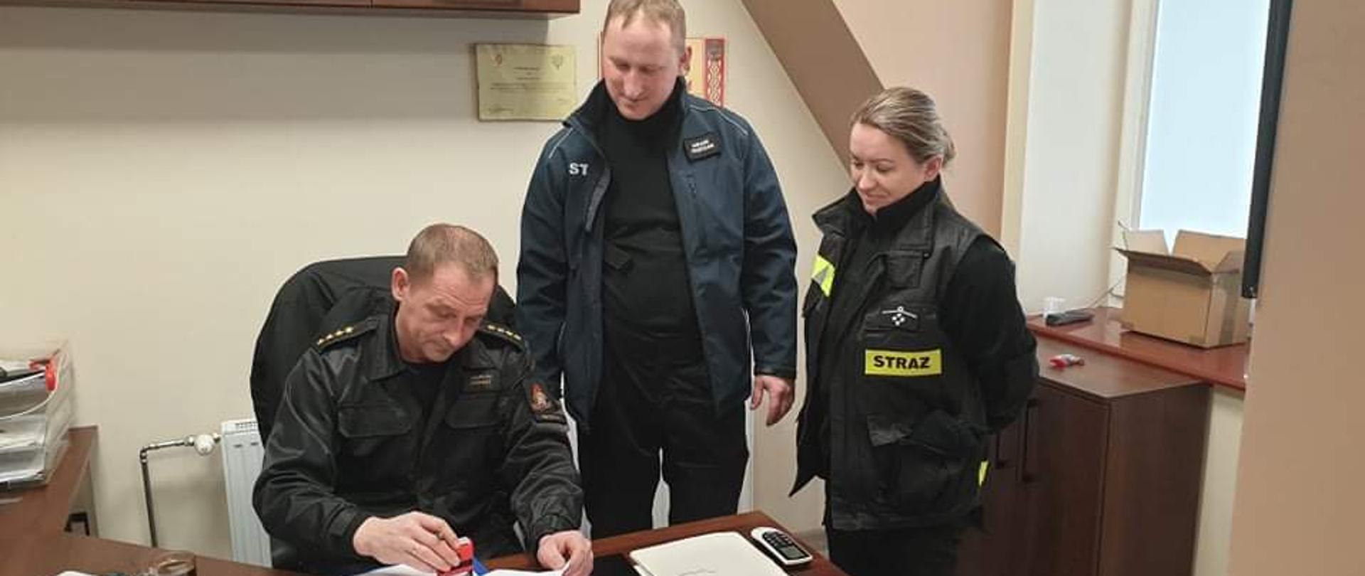 Na zdjęciu widać siedzącego na krześle strażaka z Państwowej Straży Pożarnej z czterema gwiazdkami na pagonach. Strażak podpisuje dokumenty. Obok stoi jeden strażak z ochotniczej straży pożarnej ubrany w kurtkę z częściowo widocznym napisem straż. Obok niego stoi strażaczka ubrana w ciemny strój i kamizelkę z napisem straż. Zdjęcie zrobione jest w pomieszczeniu. Na biurku leżą pisma.