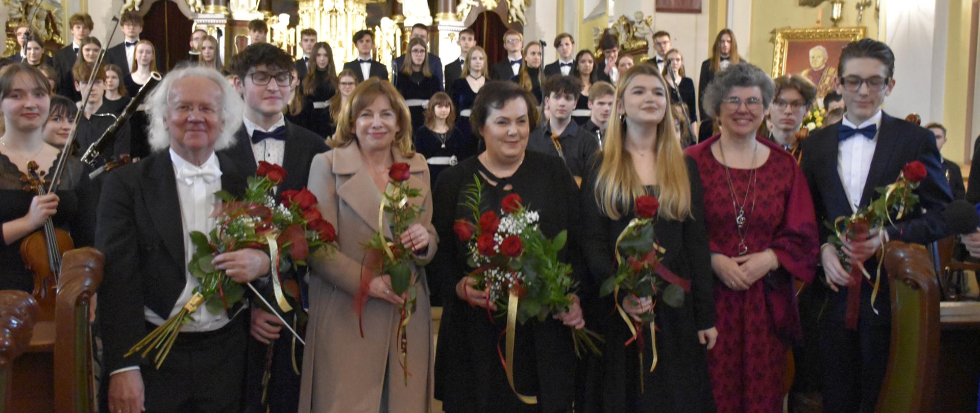 na tle kościelnego ołtarza obraz Matki Bożej, złote figury, na pierwszym planie grupa osób w pierwszym rzędzie osoby trzymają czerwone róże