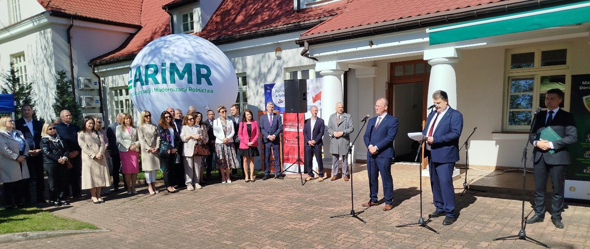 Grupa osób zgromadzona przed budynkiem z czerwonym dachem podczas oficjalnego wydarzenia. Kilku mężczyzn przemawia przy mikrofonach.