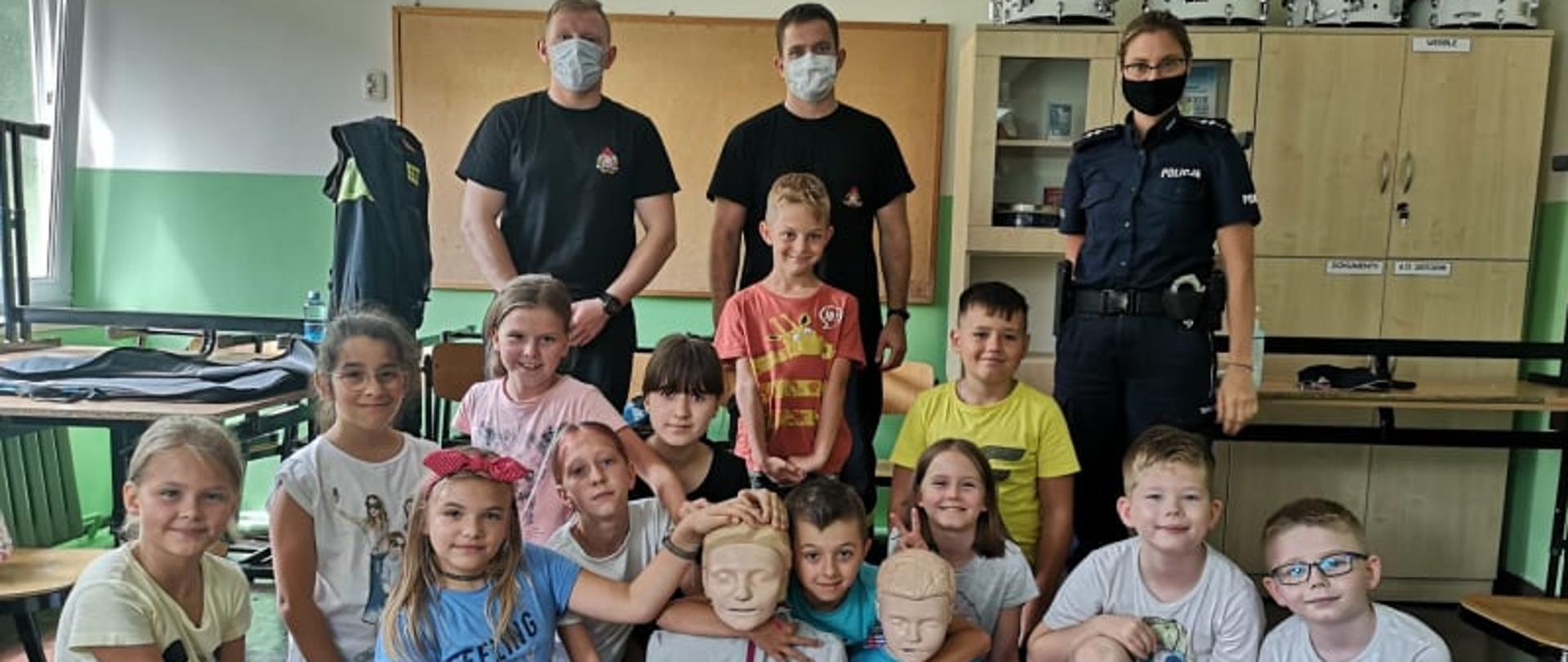 Strażacy na półkoloniach. Sala lekcyjna w Szkole Podstawowej nr 4 w Rawiczu. Dwanaścioro dzieci, uczestników półkolonii klęczy na kocu w półokręgu. Na środku dwa fantomy trzymane przez dzieci. Z tyłu klęczących stoi dwóch strażaków oraz policjantka.