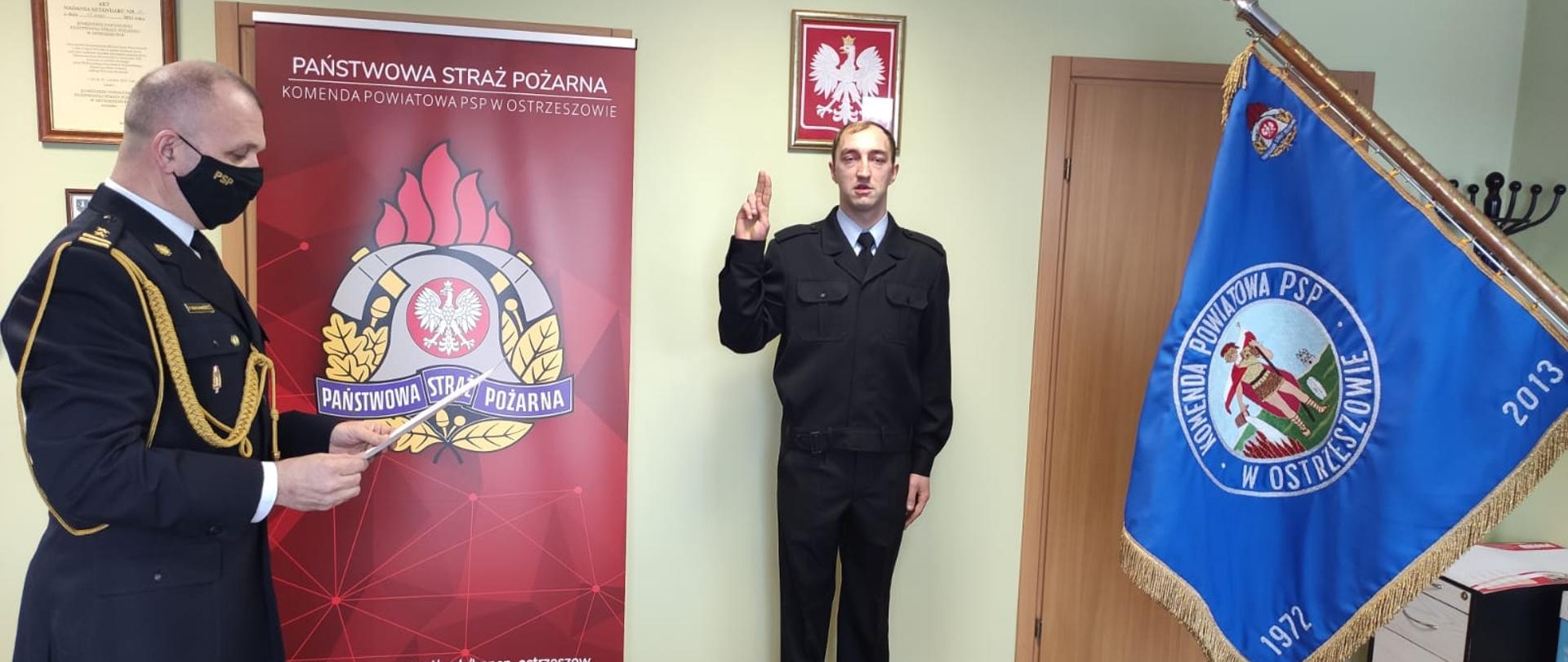 Zdjęcie przedstawia ślubowanie strażaka, który składa przysięgę zgodnie z ceremoniałem w obecności sztandaru Państwowej Straży Pożarnej w Ostrzeszowie. Po lewej stronie ślubowaniu przewodzi Komendant Powiatowy Państwowej Straży Pożarnej w Ostrzeszowie. Zdjęcie wykonane w pomieszczeniu sali konferencyjnej.