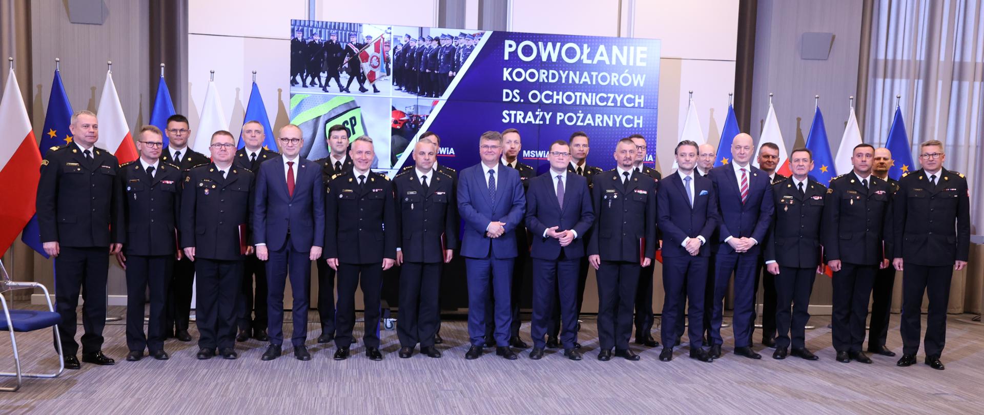 Zastępcy komendantów wojewódzkich PSP w granatowych mundurach wśród nich z pracownicy MSWiA. Za ich plecami flaga Polski i Unii Europejskiej