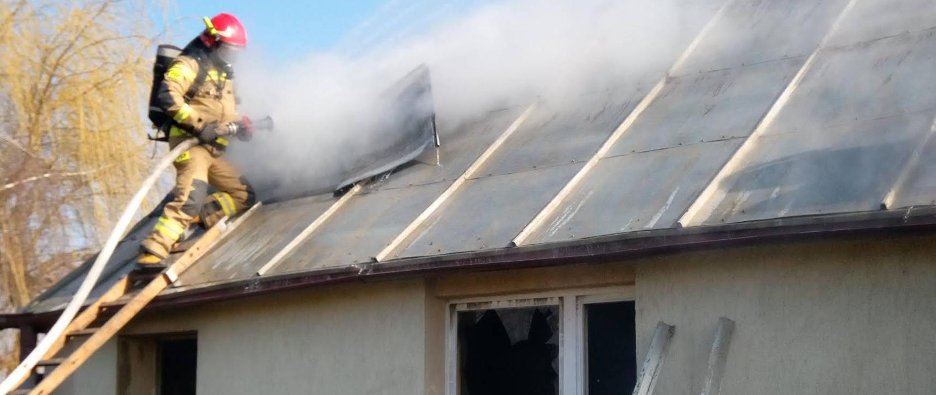 Strażak na drabinie opartej o palący się budynek, budynek parterowy, kryty blachą.