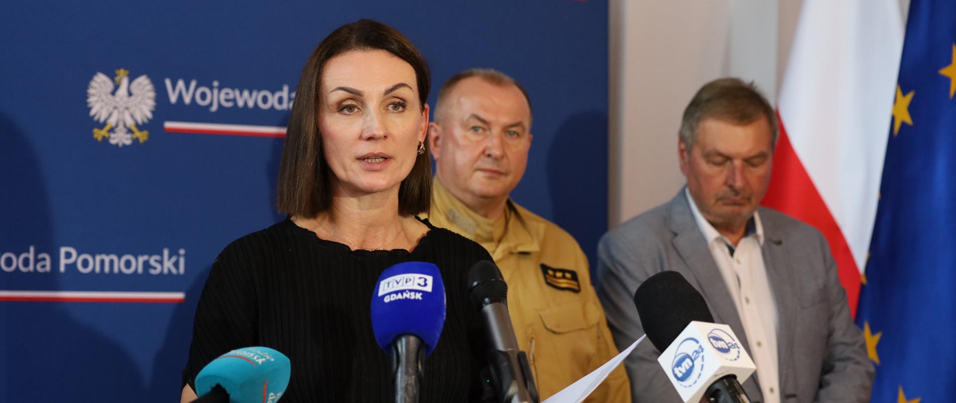 kobieta (wojewoda) udziela odpowiedzi dziennikarzom, przed nią mikrofony