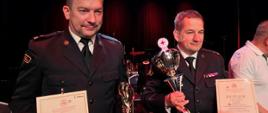 Dwaj mężczyźni w mundurach prezentują dyplomy i trofea, stojąc na scenie podczas uroczystości. W tle widać fragment instrumentów muzycznych i kolejną osobę.