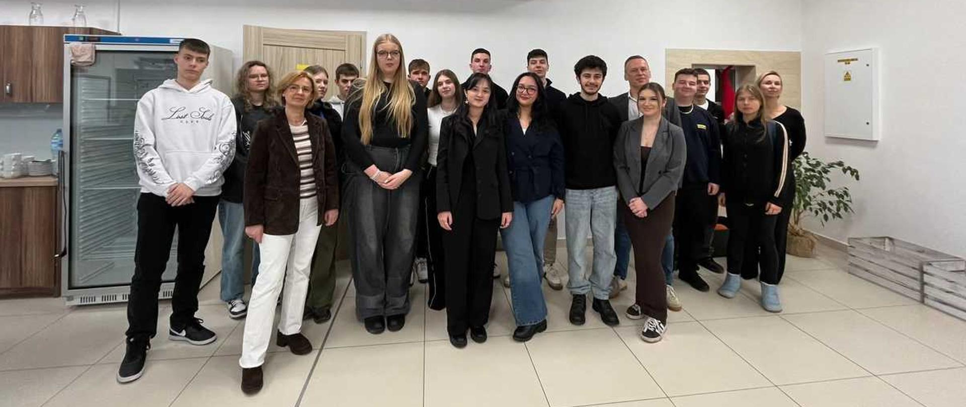 Zdjęcie przedstawia grupę kilkunastu osób, młodzieży i dorosłych, stojących razem w pomieszczeniu przypominającym pokój socjalny lub biuro, z kafelkami na podłodze i podwieszanym sufitem z oświetleniem panelowym.