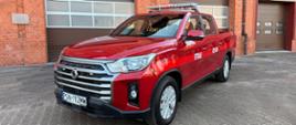 Czerwony pickup straży pożarnej zaparkowany przed budynkiem remizy z ceglanej elewacji i zamkniętymi bramami garażowymi; na dachu pojazdu znajduje się belka świetlna, na drzwiach widoczny napis „STRAŻ”.