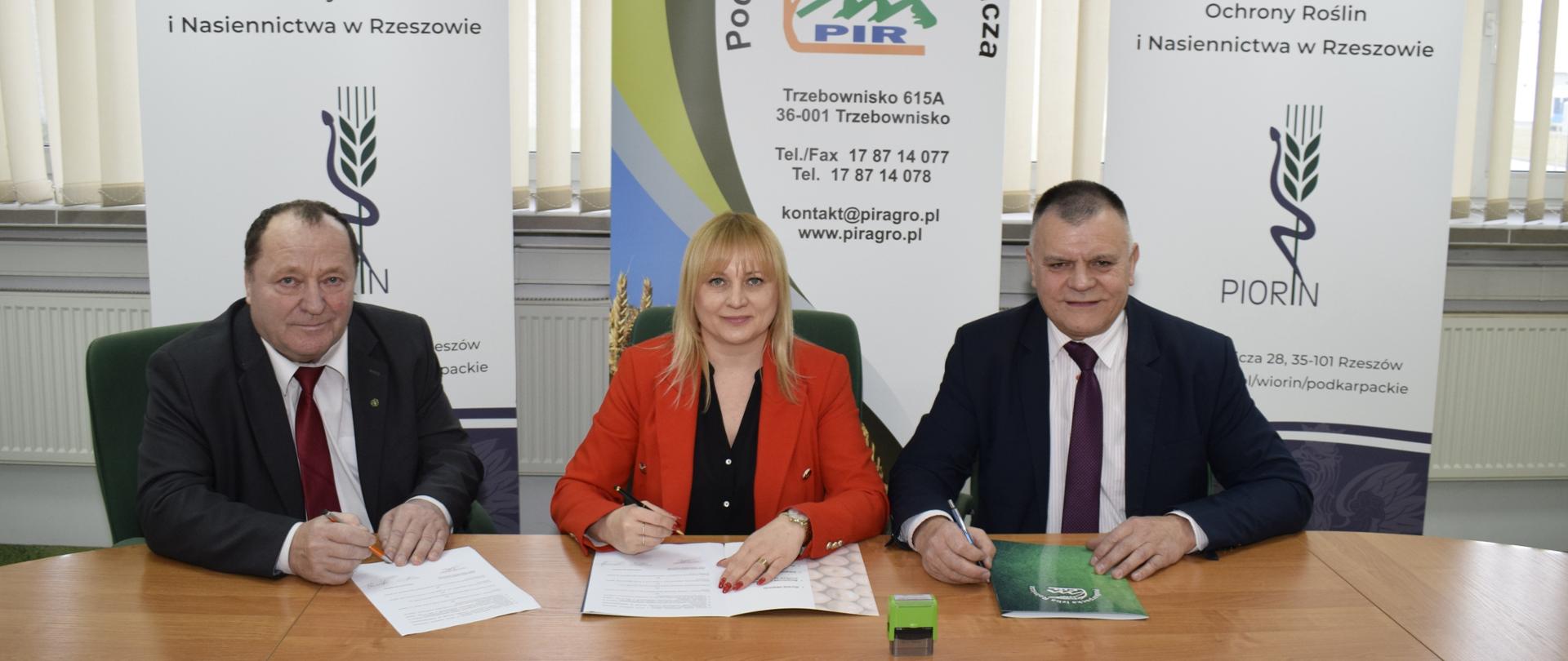 Trzy uśmiechnięte osoby siedzące przy stole konferencyjnym. Dwóch mężczyzn po zewnętrznych stronach ubranych jest w czarne garnitury białe koszule i krawaty, kobieta w środku ma na sobie czerwoną marynarkę i czarną koszulę. Wszyscy troje trzymają w dłoniach długopisy a przed nimi na stole znajdują się dokumenty. Za plecami siedzących znajdują się banery instytucji przez nich reprezentowanych.