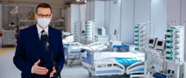 Na pierwszym planie Premier Mateusz Morawiecki stoi przy mikrofonie w sali szpitalnej w Szpitalu Tymczasowym w Krakowie. W tle puste łóżka i sprzęt medyczny.