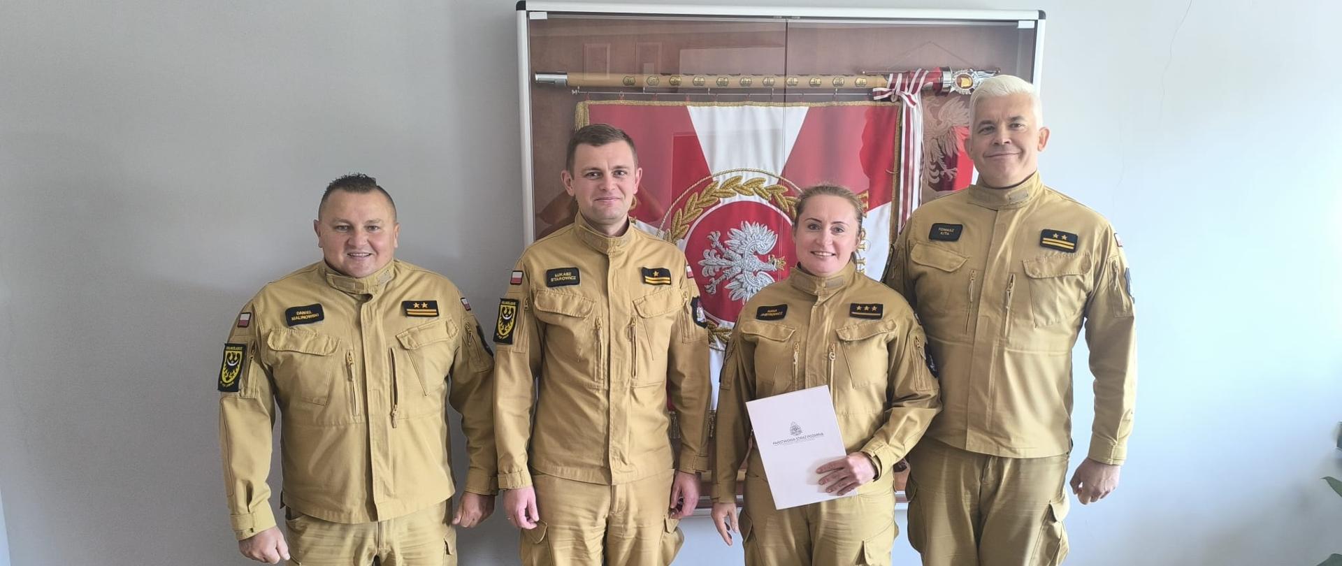 Grupa czterech strażaków w piaskowych mundurach stoi w pomieszczeniu przed gablotom ze sztandarem; jedna z osób trzyma w ręku akt mianowania na wyższe stanowisko.